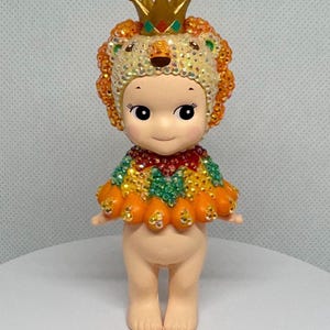 May include: A small, decorative figurine of a cherubic character wearing a jeweled lion headdress and cape. The headdress is gold with a red and green jewel. The cape is orange, green, and yellow. The figurine has a neutral skin tone.