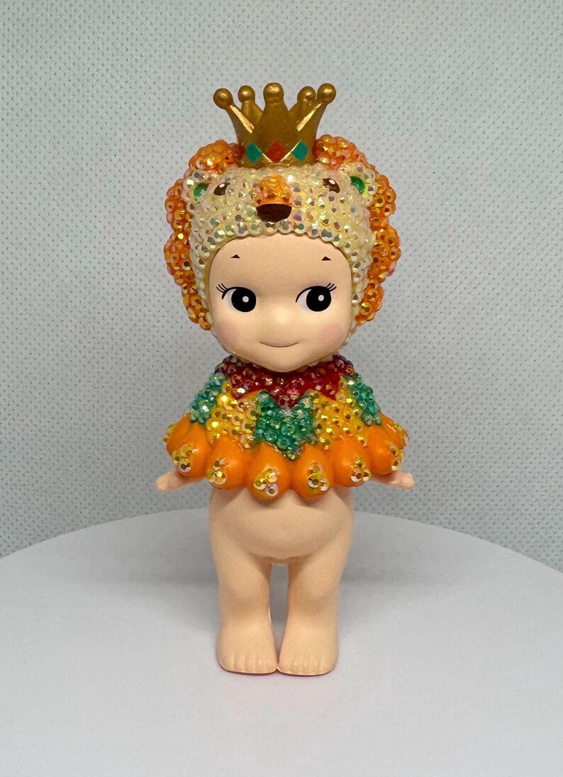 May include: A small, decorative figurine of a cherubic character wearing a jeweled lion headdress and cape. The headdress is gold with a red and green jewel. The cape is orange, green, and yellow. The figurine has a neutral skin tone.