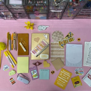 May include: A collection of stationery items including a gold notebook, yellow pens, a pom-pom pen, sticky notes, paper clips, stickers, a small pouch, a notepad with a book stack illustration, and a Swiss Miss hot chocolate packet on a pink surface.