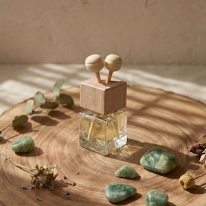 Puede incluir: Un frasco de vidrio cuadrado que contiene un líquido claro, rematado con una tapa de madera en forma de cubo y dos palitos de madera con extremos esféricos. El frasco está rodeado de varias piedras verdes pulidas y elementos botánicos secos.