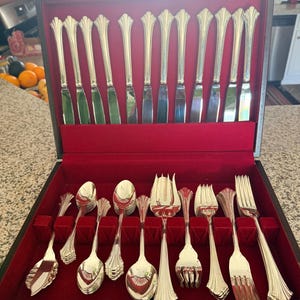 May include: A silver-colored flatware set displayed in a wooden box with a red velvet interior. The set includes knives, spoons, forks, and serving utensils. The handles have a decorative, ornate design.
