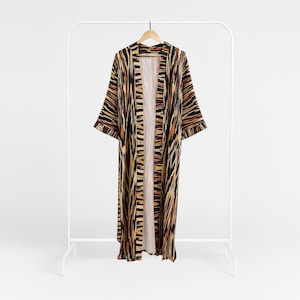 Puede incluir: Larga bata kimono con frente abierto, con un llamativo estampado abstracto. La bata presenta una mezcla de rayas negras, marrones y naranjas, creando un efecto visual impactante. Cuelga de un perchero blanco, mostrando su diseño fluido y su tejido ligero.