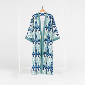 Puede incluir: Una bata kimono larga y abierta con un estampado azul, turquesa y blanco. La bata está colgada en un perchero blanco. Las mangas son cortas y anchas, y el ribete es turquesa. La bata tiene una borla al final de las cintas.