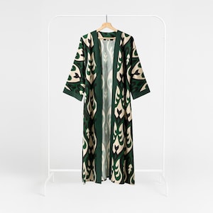 Puede incluir: Bata larga con frente abierto con un estampado verde, crema y negro. La bata está colgada en un perchero blanco con una percha de madera. El estampado presenta formas estilizadas de hojas y flores.