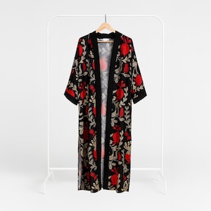 Puede incluir: Una bata tipo kimono negra con un llamativo estampado floral de granadas rojas y hojas doradas. La bata cuelga de un perchero blanco, revelando su frente abierto y mangas cortas. El diseño crea un contraste llamativo sobre la tela negra.