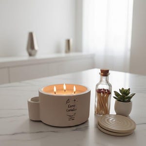 Puede incluir: Una vela beige de tres mechas, con el texto "Luno Candles", está encendida sobre una superficie de mármol blanco. Una pequeña botella de vidrio llena de cerillas, una pequeña planta suculenta en maceta y una tapa de cerámica están cerca.