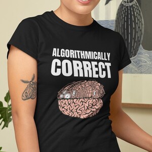 May include: Black t-shirt with the text "ALGORITHMICALLY CORRECT" in white. The graphic below the text depicts a brain with gears and circuits. The person in the image is wearing black pants and has a tattoo of a moth on their arm.