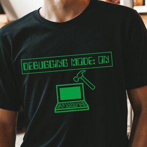 May include: Black t-shirt with a green graphic that reads "DEBUGGING MODE: ON". The design includes a laptop and a hammer. The shirt is made of a soft material.