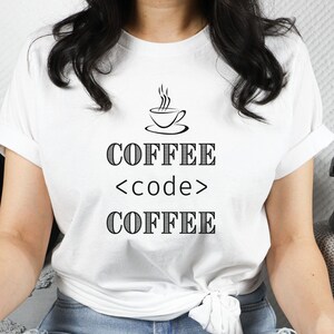 May include: White t-shirt with the text "COFFEE <code > COFFEE" in black, along with a small coffee cup graphic. The shirt is worn with blue jeans. A black hat and skull are in the background.