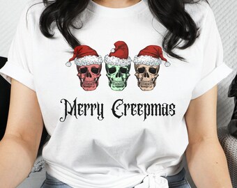 May include: White t-shirt featuring three colorful skulls wearing Santa hats, with the text "Merry Creepmas" in a gothic font. The skulls are pink, green, and tan.