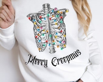 May include: White sweatshirt featuring a detailed illustration of a rib cage adorned with colorful Christmas lights. The phrase "Merry Creepmas" is printed in a gothic font below the rib cage design. A festive and spooky holiday theme.