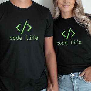 May include: Black t-shirts with the text "code life" in green, along with a green code symbol. The shirts are worn by two people, with the focus on the shirts and text. The people are wearing blue jeans.
