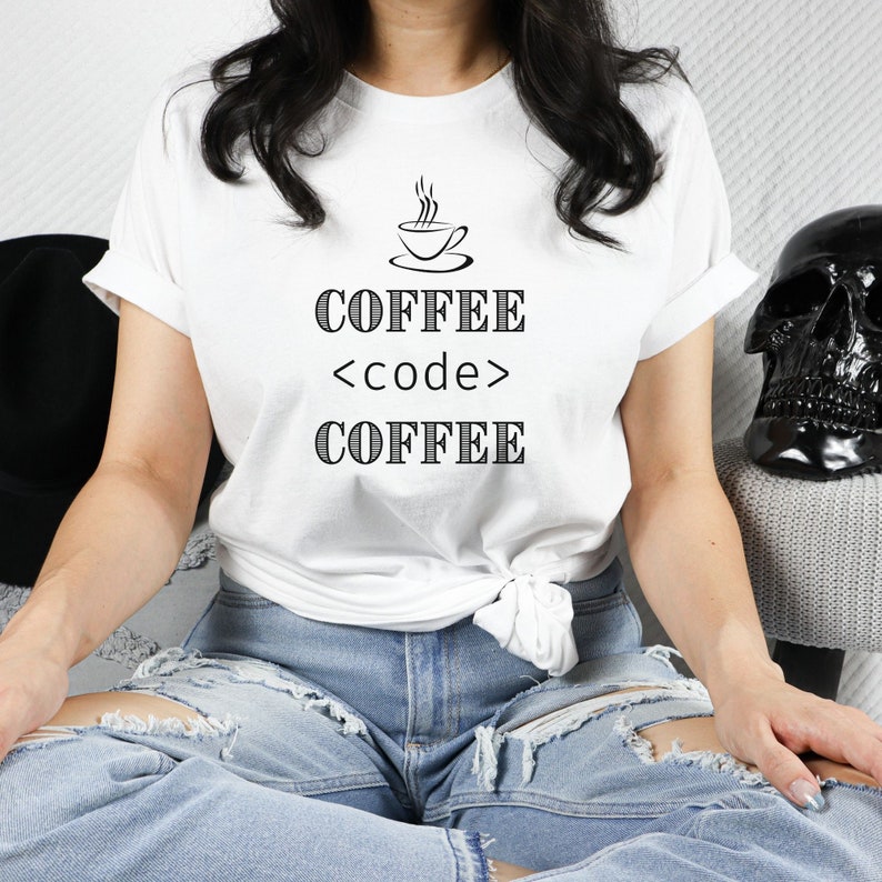 May include: White t-shirt with the text "COFFEE <code > COFFEE" in black, along with a small coffee cup graphic. The shirt is worn with blue jeans. A black hat and skull are in the background.