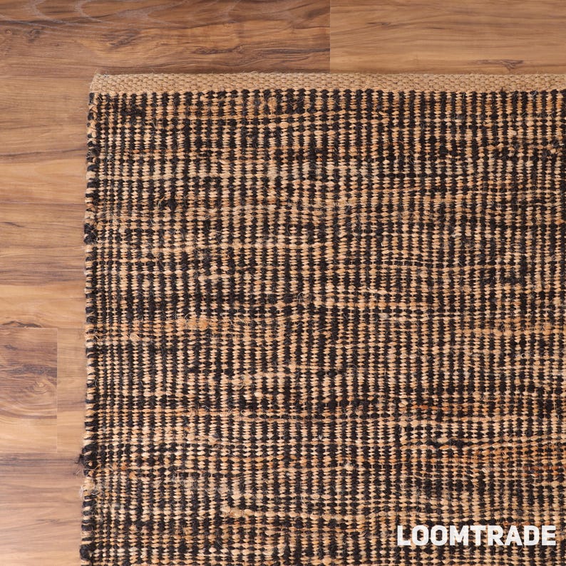 May include: A rectangular area rug featuring a woven texture with alternating vertical stripes of dark brown and natural tan fibers. The rug has a thick, braided border along the top edge and the text LOOMTRADE is printed in white in the bottom corner.