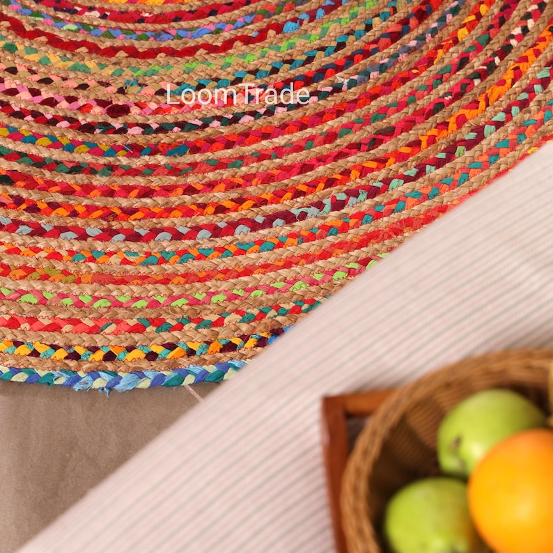 May include: Close-up of a round, woven jute rug with concentric bands of color. The rug features a mix of red, orange, green, and blue hues. The text "Loom Trade" is visible on the rug. A basket of fruit is in the foreground.