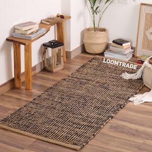 May include: A rectangular woven area rug featuring a textured pattern of alternating natural tan and black fibers. The rug has a flat-weave construction with visible horizontal and vertical interlacing. The word LOOMTRADE is printed in white.