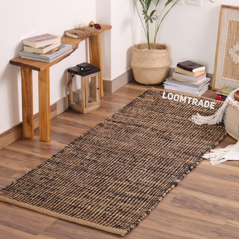 May include: A rectangular woven area rug featuring a textured pattern of alternating natural tan and black fibers. The rug has a flat-weave construction with visible horizontal and vertical interlacing. The word LOOMTRADE is printed in white.