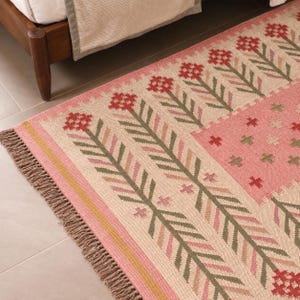 May include: A woven area rug featuring a cream-colored base with repeating vertical rows of stylized red flowers and green leaves. The rug includes a solid pink rectangular section with small cross motifs and a fringed edge in a natural tan color.