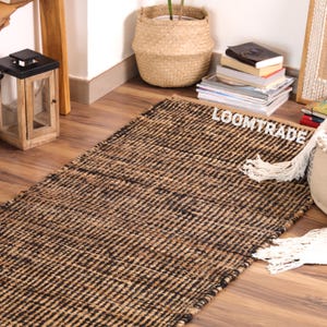 May include: A rectangular area rug featuring a woven pattern of alternating natural tan and black fibers. The rug has a textured surface and includes white fringe detailing on one side. The word LOOMTRADE is printed in white on the rug surface.