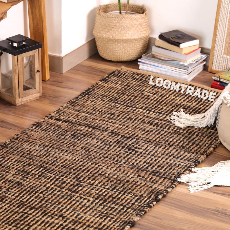 May include: A rectangular area rug featuring a woven pattern of alternating natural tan and black fibers. The rug has a textured surface and includes white fringe detailing on one side. The word LOOMTRADE is printed in white on the rug surface.
