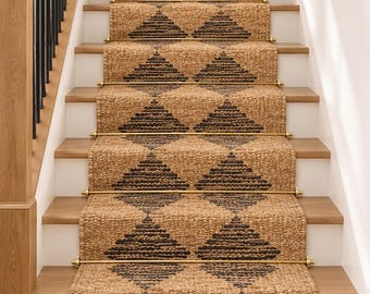 May include: A woven natural fiber stair runner featuring a repeating dark brown diamond pattern on a tan background. The runner is secured to the wooden stairs with metallic gold rods and displays the text LOOMTRADE on the top step.