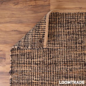 May include: A rectangular area rug featuring a woven pattern of alternating black and natural tan fibers. The rug has a thick, braided border edge and is folded at one corner to reveal the textured construction and the text LOOMTRADE.