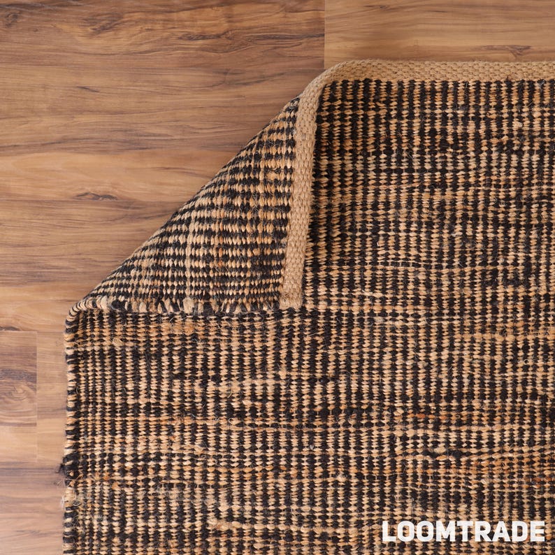 May include: A rectangular area rug featuring a woven pattern of alternating black and natural tan fibers. The rug has a thick, braided border edge and is folded at one corner to reveal the textured construction and the text LOOMTRADE.
