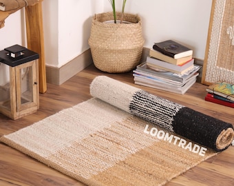 May include: A rectangular woven rug featuring a color-blocked design with sections of cream, tan, and a black and white striped pattern. The rug is partially rolled, revealing a natural fiber texture and the white text LOOMTRADE printed on it.