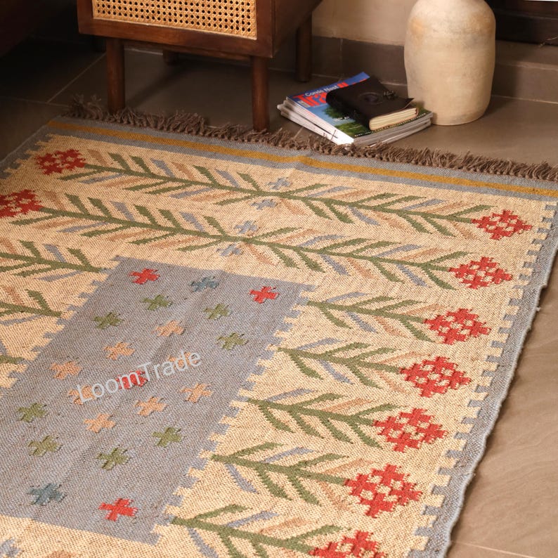 May include: A rectangular woven rug featuring a natural beige base with a repeating pattern of green leafy stems and red floral motifs. A central gray rectangular panel displays small cross-shaped icons in red, green, and blue, with the text LoomTrade.