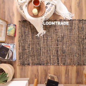 May include: A rectangular area rug featuring a woven texture with alternating vertical stripes of natural tan and dark brown fibers. The rug displays the white text LOOMTRADE in the upper center area, set against a backdrop of home decor items.
