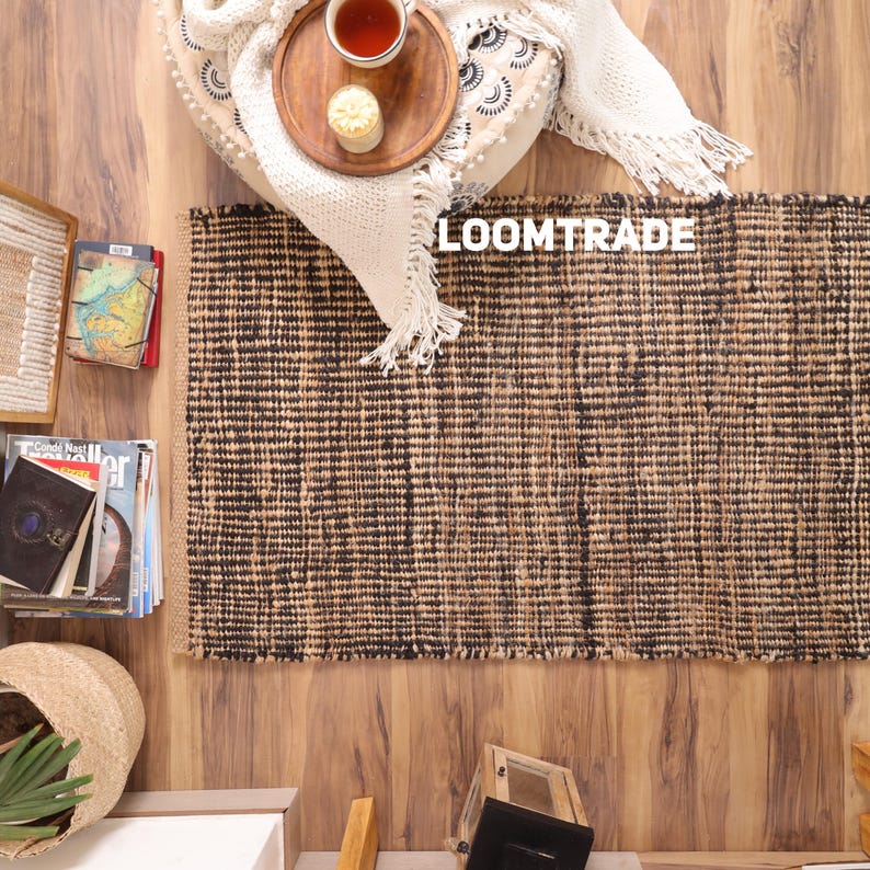 May include: A rectangular area rug featuring a woven texture with alternating vertical stripes of natural tan and dark brown fibers. The rug displays the white text LOOMTRADE in the upper center area, set against a backdrop of home decor items.