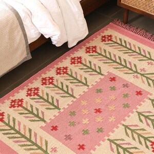 May include: A rectangular woven area rug featuring a cream-colored base with a repeating pattern of red flowers on green stems. The rug includes a central pink rectangular panel decorated with small red, green, and beige cross-stitch motifs.