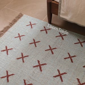 May include: A light blue rug with a repeating pattern of brown crosses. The rug has a fringed edge and the text "LoomTrade" is visible on the rug's surface. The rug is near a wooden bed frame.
