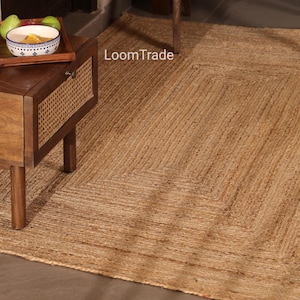 Peut inclure: Un tapis rectangulaire en jute avec un motif tissé. Le tapis est de couleur beige naturel et a une bordure définie. Le texte "LoomTrade" est visible sur le tapis.