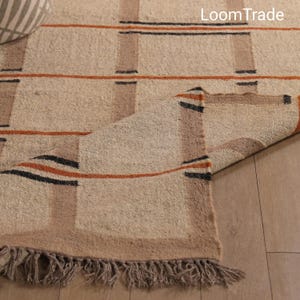 Könnte beinhalten: Beiger gewebter Teppich mit einem geometrischen Muster aus braunen, schwarzen und orangefarbenen Streifen und Quadraten. Der Teppich hat einen Fransenrand und den Text "LoomTrade" in der oberen rechten Ecke.