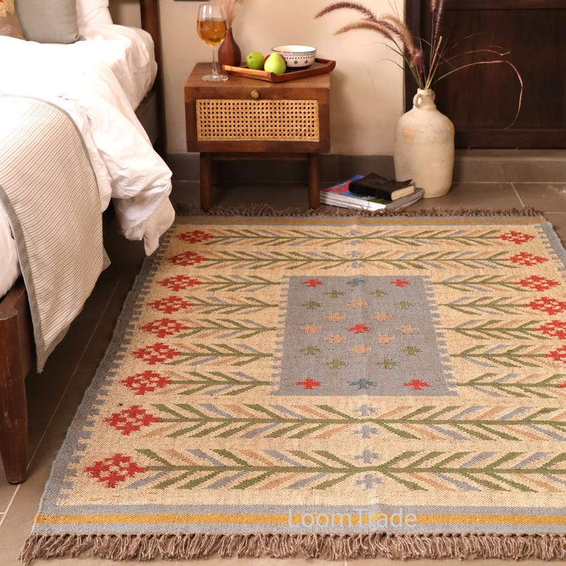 May include: A rectangular area rug featuring a beige base with a repeating pattern of red floral motifs and green stems. The center contains a light blue rectangular panel with small red and green cross-stitch accents. The rug has fringed edges.