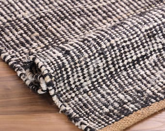 May include: A woven area rug featuring a textured pattern of alternating black and off-white fibers. The rug is partially folded, revealing a thick, natural tan jute border along the edge. The text LOOMTRADE is visible in the top right corner.