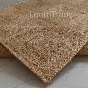 May include: A natural jute rug with a geometric design. The rug is a warm tan colour and has a woven texture. The corner of the rug is folded over, revealing the underside. The text "LoomTrade" is visible on the rug.
