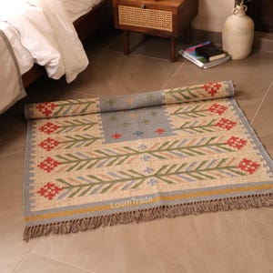 May include: A rectangular woven rug featuring a beige base with a repeating pattern of red and green floral motifs. The rug has a blue central panel with small cross designs, a yellow border stripe, and fringed edges. The text LoomTrade is visible.