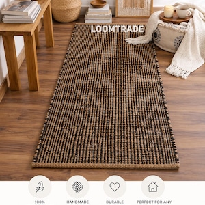 May include: A rectangular runner rug featuring a woven pattern of alternating natural tan and black jute fibers. The rug has a textured surface and a consistent weave throughout its length. The text LOOMTRADE is visible on the rug surface.