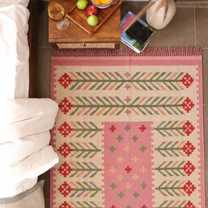May include: A rectangular area rug featuring a beige base with a central pink panel and a repeating pattern of red, green, and purple stylized floral motifs. The rug has a fringed edge and a woven texture, displaying a symmetrical design.