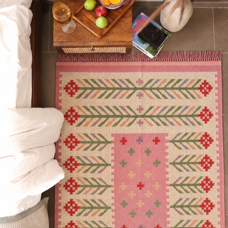 May include: A rectangular area rug featuring a beige base with a central pink panel and a repeating pattern of red, green, and purple stylized floral motifs. The rug has a fringed edge and a woven texture, displaying a symmetrical design.