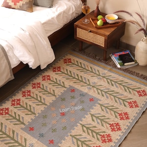 May include: A rectangular woven rug featuring a beige base with a central light blue panel. The design includes repeating floral motifs in red and green, with the text LoomTrade printed in the center. The rug has a fringed edge on the short sides.