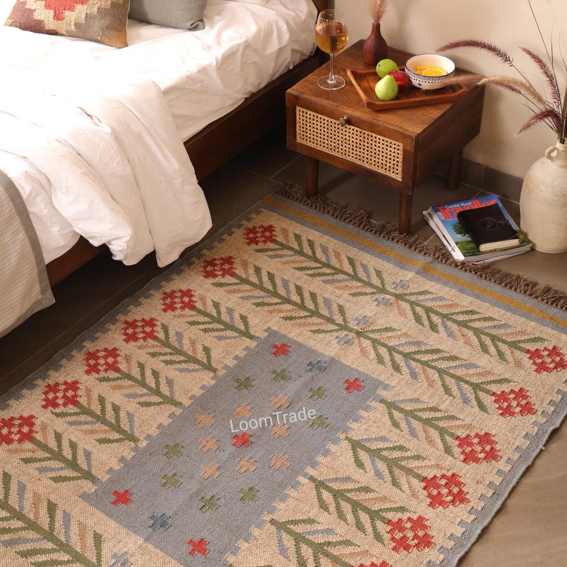 May include: A rectangular woven rug featuring a beige base with a central light blue panel. The design includes repeating floral motifs in red and green, with the text LoomTrade printed in the center. The rug has a fringed edge on the short sides.