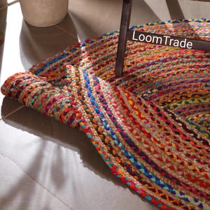 May include: Braided jute rug with vibrant colors including red, blue, orange, and green. The rug is partially rolled up, revealing the woven texture and the text "LoomTrade". This decorative rug adds a bohemian touch to any space.