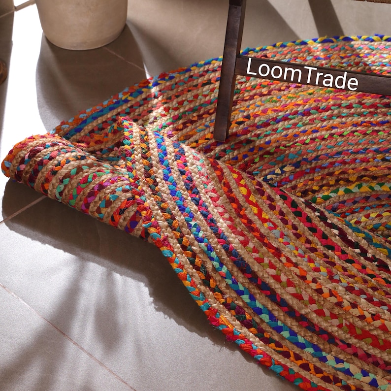 May include: Braided jute rug with vibrant colors including red, blue, orange, and green. The rug is partially rolled up, revealing the woven texture and the text "LoomTrade". This decorative rug adds a bohemian touch to any space.