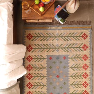 May include: A rectangular woven rug featuring a natural beige base with a central light blue panel. The design includes repeating red and green geometric floral motifs and small cross patterns. The word LoomTrade is printed in white text.