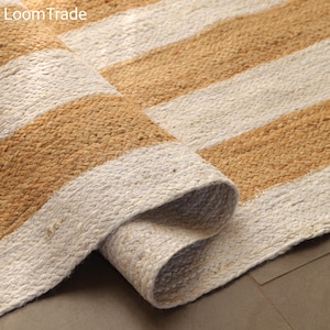 May include: A rolled-up jute rug with a striped design. The rug features alternating bands of beige and off-white, creating a textured pattern. The edge of the rug is visible, showcasing the woven construction. The brand name "LoomTrade" is in the top left corner.