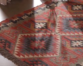 May include: A rectangular, patterned rug with a geometric design in shades of red, gray, and beige. The rug has a fringed edge and the text "LoomTrade" is visible in the upper right corner. The rug's design features repeating diamond and square patterns.
