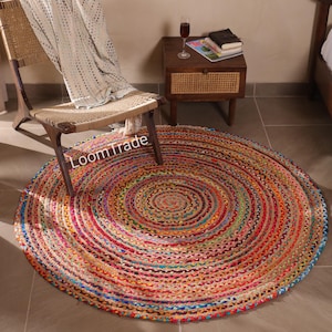 May include: A round, multi-colored jute rug with concentric circles of red, orange, blue, and green. The rug is on a tiled floor, with a wooden chair and side table in the background. The text "LoomTrade" is visible on the rug.